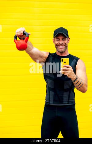 Positive smiling muscular male athlete in black activewear lifting kettlebell and using mobile phone while standing over yellow wall Stock Photo