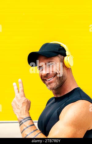 Portrait of smiling handsome male athlete in black cap and activewear showing peace sign and listening music over headphones against yellow wall Stock Photo