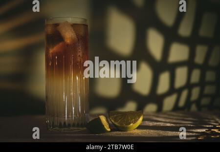 Dark and Stormy cocktail with ice and lime wedges on white table cloth in front of a green dappled background/wall Stock Photo