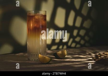 Dark and Stormy highball cocktail with ice and lime wedges on white table cloth in front of a green dappled background Stock Photo