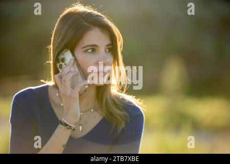 Relaxed woman talking on the phone outdoors against sunset backlight. Technology and communication concept. Stock Photo