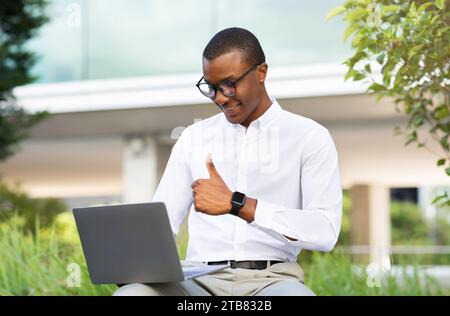 Black Businessman Using Laptop Outdoors, Making Video Call And Showing Thumb Up Stock Photo