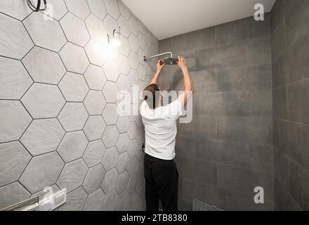 Back view of man standing by the wall with ceramic tile and installing high pressure bath showerhead at home. Male worker fixing bathroom showerhead while working on bathroom renovation. Stock Photo