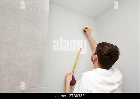 Male worker using tape measure and pencil while checking wall level before repair works. Man measuring white wall and drawing line. Home renovation concept. Stock Photo