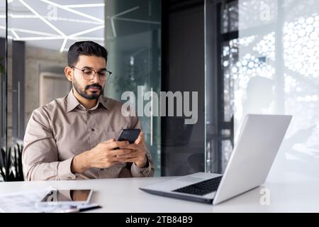 Serious concentrated businessman at the workplace inside the office, the man holds a smartphone in his hands, a worker with a laptop browses the Inter Stock Photo