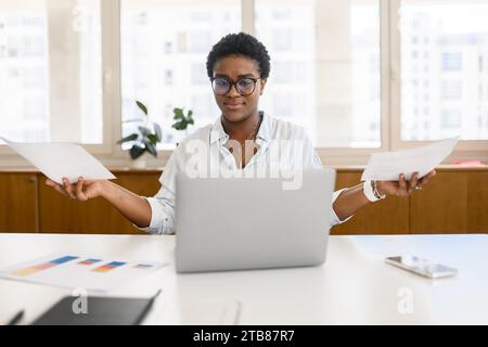 Worried puzzled African-American female office employee sitting inside at the desk and looking through working papers, contracts, confused woman found mistake or inconsistency in annual report Stock Photo