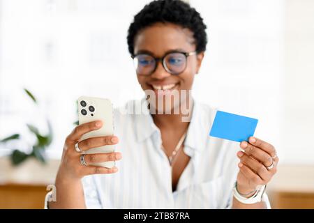 Excited smiling African-American woman is using a smartphone and a credit card for shopping online, happy lady is ordering food online, paying for long-awaited purchase on the mobile phone. E-banking Stock Photo