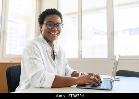 Side view at stylish african american business woman with short haircut in casual outfit sitting in the workplace and using laptop, smiling at camera, posing alone at cozy office Stock Photo