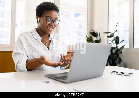 Friendly smiling african american young businesswomanwith short haircut using laptop for video call, talking online and gesturing. Pleasant multiracial female employee holding video call with client Stock Photo