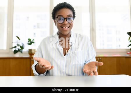 Attractive energetic African-American female colleague, tutor, coach talking online during video call, sitting at the desk, looking at the camera and talking with gestures. Virtual meeting, conference Stock Photo