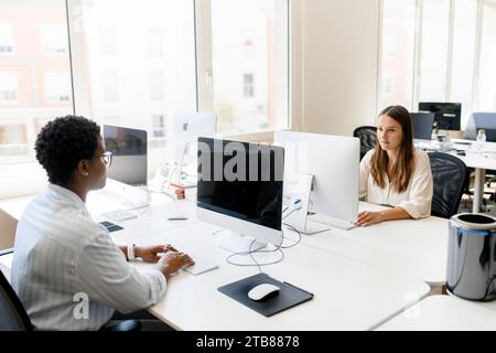 Two female colleagues sitting at the desk in modern open space office, smart and concentrated women working on the computers, office stuff on the workplace, enjoying friendly working atmosphere Stock Photo