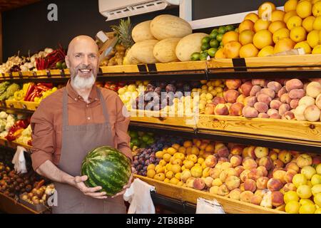 joyous good looking seller with beard posing with fresh watermelon in hands and smiling at camera Stock Photo