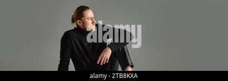 banner of bearded man in turtleneck sweater and black leather pants sitting on grey background Stock Photo
