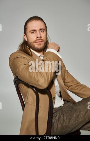 good looking bearded businessman in beige formal attire sitting on wooden chair on grey background Stock Photo