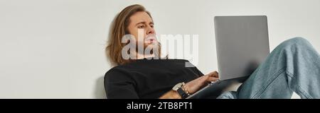 handsome man with long hair and beard leaning on wall and using laptop on grey backdrop, banner Stock Photo