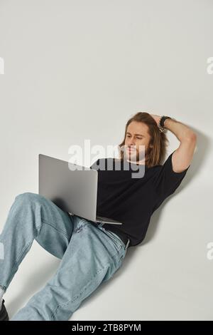 handsome man with long hair and beard leaning on white wall and using laptop on grey backdrop Stock Photo
