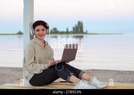Attractive young freelancer working on a beach with headphones and laptop smiling at the camera Stock Photo