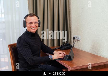 Smiling freelance asian man wearing eyeglasses and headphones working at home and using tablet computer and looking at the camera Stock Photo