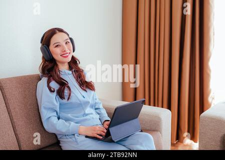 Closeup of smiling freelance asian woman working at home wearing headphones and using tablet computer and looking at the camera Stock Photo