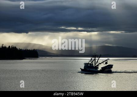 Fishing boat motors through the Inside Passage with sunlight shining through dark clouds in the distance; Alaska, United States of America Stock Photo