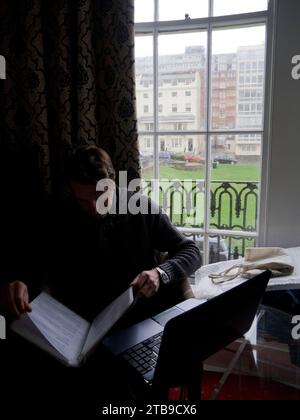 Man sitting by a window with laptop reading documents in a hotel room in Brighton on a rainy day; Brighton, East Sussex, England, United Kingdom Stock Photo