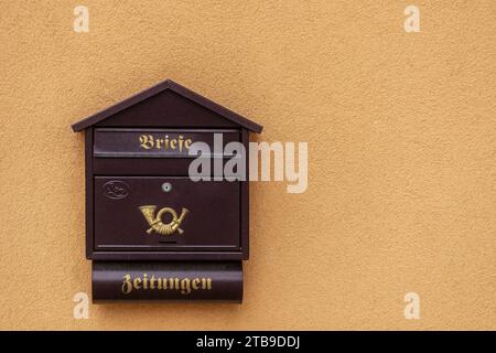 Brown mailbox with an emblem against a background of a plastered orange wall with gold lettering. Translation: "Letters", "Newspapers". Stock Photo