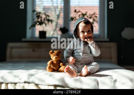 Little boy in gray pajamas sitting on the bed with his favorite toy teddy bear. Happy childhood concept Stock Photo