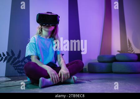 School aged girl child in VR glasses sitting on the floor, having an immersive experience through interaction with virtual reality. Stock Photo