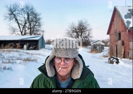 Man smoking a cigarette stands in the snow at a farm; Dunbar, Nebraska, United States of America Stock Photo