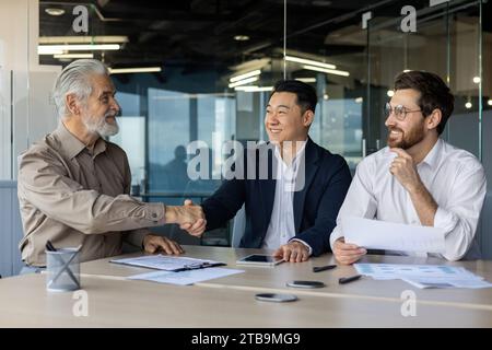 Making a business deal in the office. Three interracial men of different ages sit at a table with documents and negotiate while shaking hands. Stock Photo