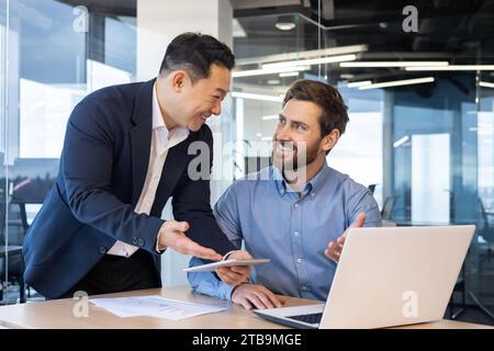 Young two interracial businessmen working in office with documents, agreement, project on tablet and laptop. Stock Photo