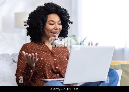 Close-up photo of a young African-American woman sitting on the couch at home and talking on a video call through a laptop, smiling, gesturing with her hands. Stock Photo