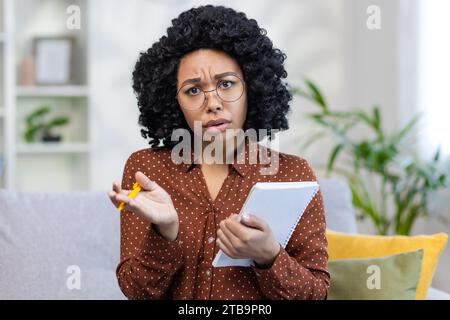 Close-up portrait of a young African American female teacher sitting at home and teaching online, holding a notebook with a pen in her hands, and talking seriously and looking at the camera. Stock Photo