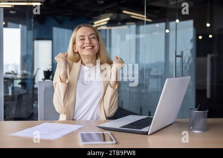 Close-up portrait of a happy business woman who is close eyes happy about success and achievement, sitting at the desk in the office and showing a victory gesture with her hands, happy for the camera. Stock Photo