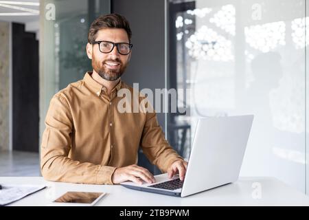 Portrait of successful man at workplace inside office, experienced smiling businessman in shirt smiling and looking at camera, programmer developing new software with laptop. Stock Photo