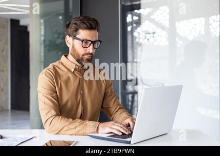 Serious concentrated thinking businessman working inside office with laptop, mature enlightened man in shirt and glasses at workplace typing on keyboard, programmer developer. Stock Photo