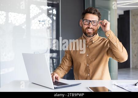 Portrait of successful man at workplace inside office, experienced smiling businessman in shirt smiling and looking at camera, programmer developing new software with laptop. Stock Photo