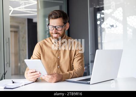 A young male programmer developer works in the office at the table at the laptop and holds a tablet in his hand. Stock Photo