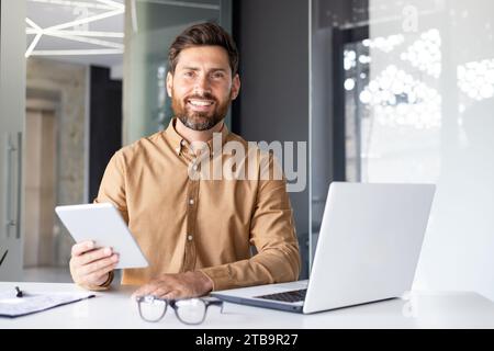 Portrait of a young male programmer working in the office at a laptop and holding a tablet in his hands, smiling and confidently looking at the camera. Stock Photo