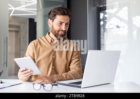 A young male programmer and developer sits in the office at the table and works on a laptop and with a tablet. Talks on a video call. Stock Photo