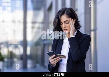Problems with the financial account. Upset young business woman standing on the street and holding her head, holding a credit card and looking at the phone. Stock Photo