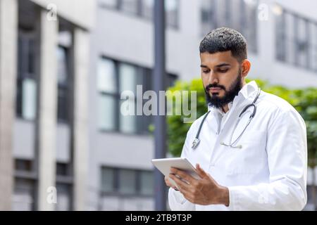 A serious young male muslim doctor is standing outside on the street near the clinic and using a tablet. Stock Photo