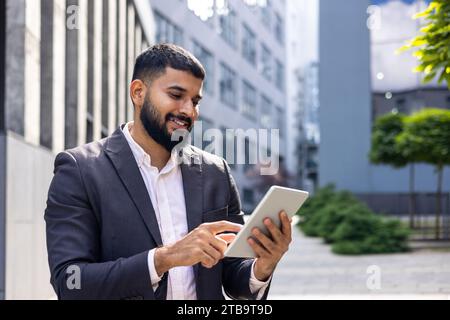 Smiling young Muslim male businessman using tablet while standing near office building, talking on video call. Stock Photo