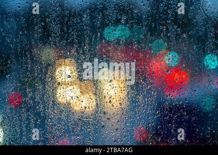 Rainy weather, road traffic, raindrops on a window pane, behind it, blurred, lights of vehicles on a rainy road, traffic lights, tail lights, headligh Stock Photo