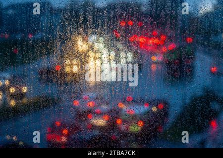 Rainy weather, road traffic, raindrops on a window pane, behind it, blurred, lights of vehicles on a rainy road, traffic lights, tail lights, headligh Stock Photo