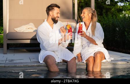 Two people in white bathrobes toasting with cocktails by the poolside, enjoying the sunset Stock Photo