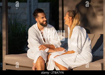 happy couple in white bathrobes sitting and talking on an outdoor bench lounger in sunlight at spa wellness hotel Stock Photo