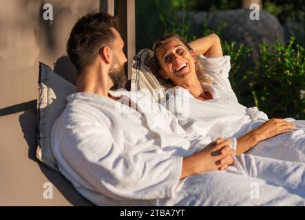 Couple in white bathrobes relaxing and laughing together on lounger in sunlight at a spa wellness hotel Stock Photo