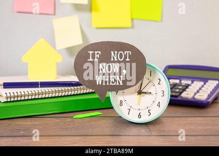Business concept. The alarm clocks have a sticker with the inscription - If Not Now, When. There are office items in the background in a blurry backgr Stock Photo