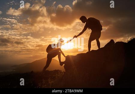 Male and female hikers climbing up mountain cliff and one of them giving helping hand. People helping and, team work concept. Stock Photo